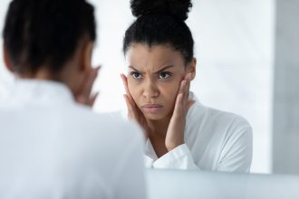 Woman looking herself in the mirror, holding her face because her face is dry.